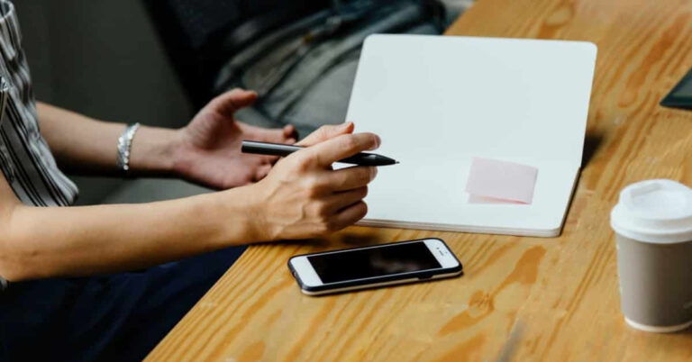Main tenant un stylo au-dessus d'un bloc-notes blanc avec un téléphone et un café