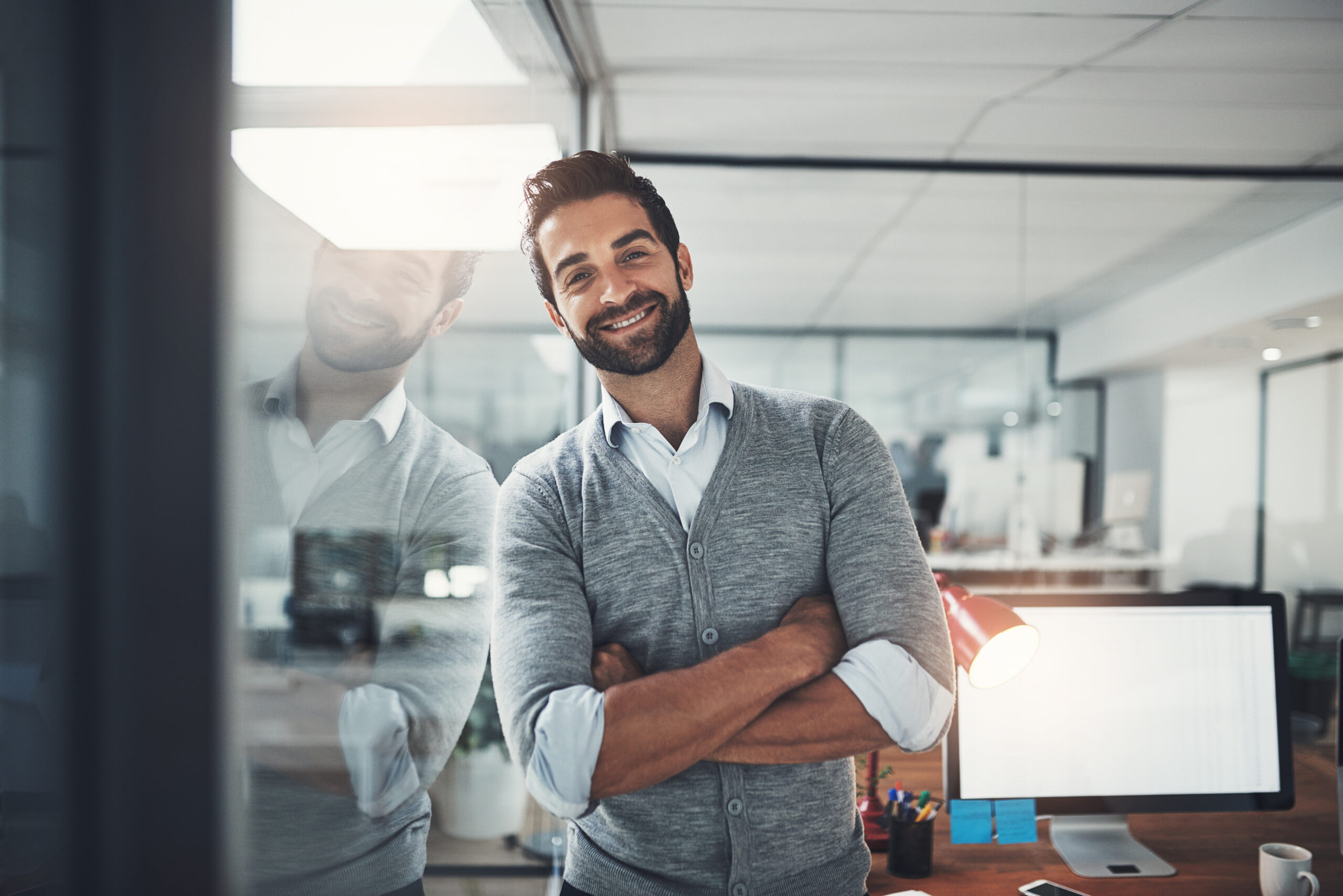 Un homme souriant, les bras croisés, se tient devant une vitre dans un bureau moderne avec un ordinateur de bureau allumé en arrière-plan.